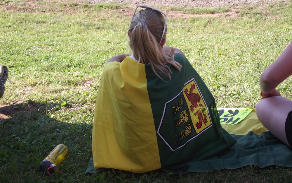 A girl is sitting on the grass with her back towards the camera. Draped around her like a cape is the Saskatchewan flag.