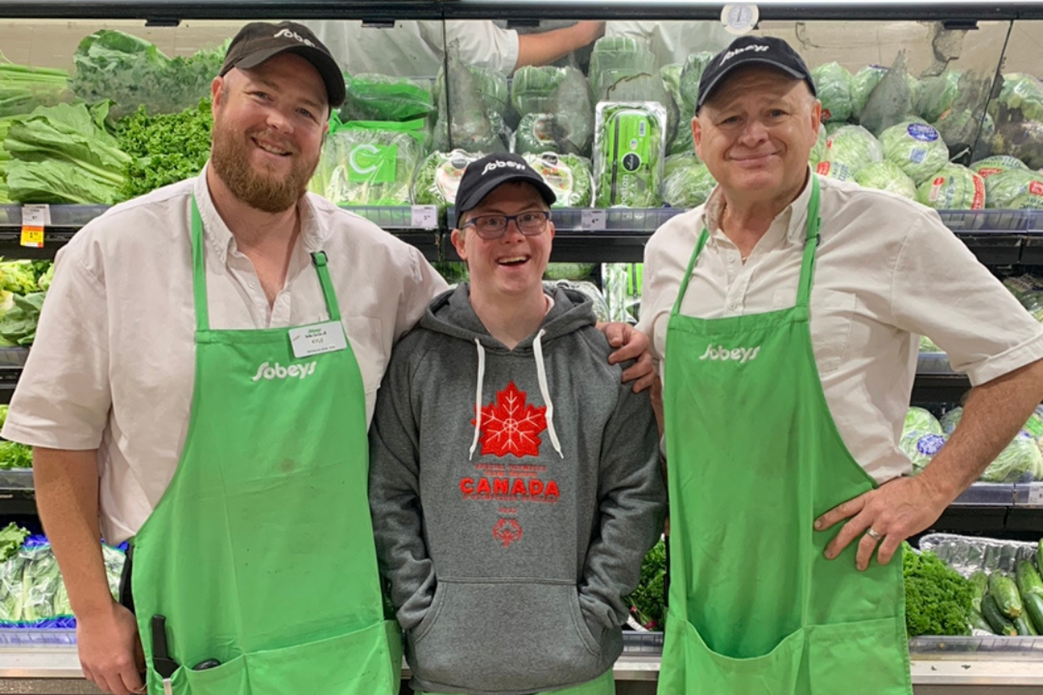 Three men standing beside each other. The two men on either end are wearing white shirts and green aprons with the Sobeys logo. The man in the middle is wearing a gret hoodie with Canada logo. All are smiling at the camera. 