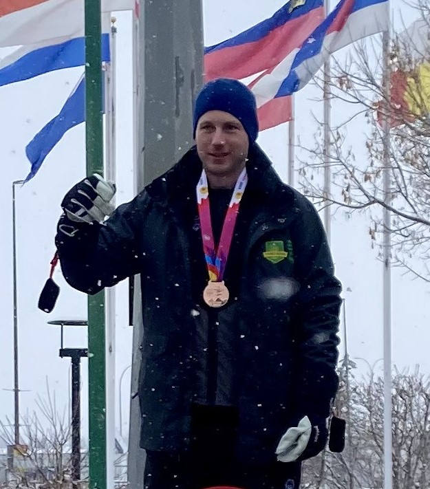A photo of Taylor, a Special Olympics Saskatchewan athlete. He is standing outside wearing a winter jacket and hat. There is snow falling around him and you can see flags waving behind him. He has a medal around his neck and putting his arm up. A photo of Taylor, a Special Olympics Saskatchewan athlete. He is standing outside wearing a winter jacket and hat. There is snow falling around him and you can see flags waving behind him. He has a medal around his neck and putting his arm up.