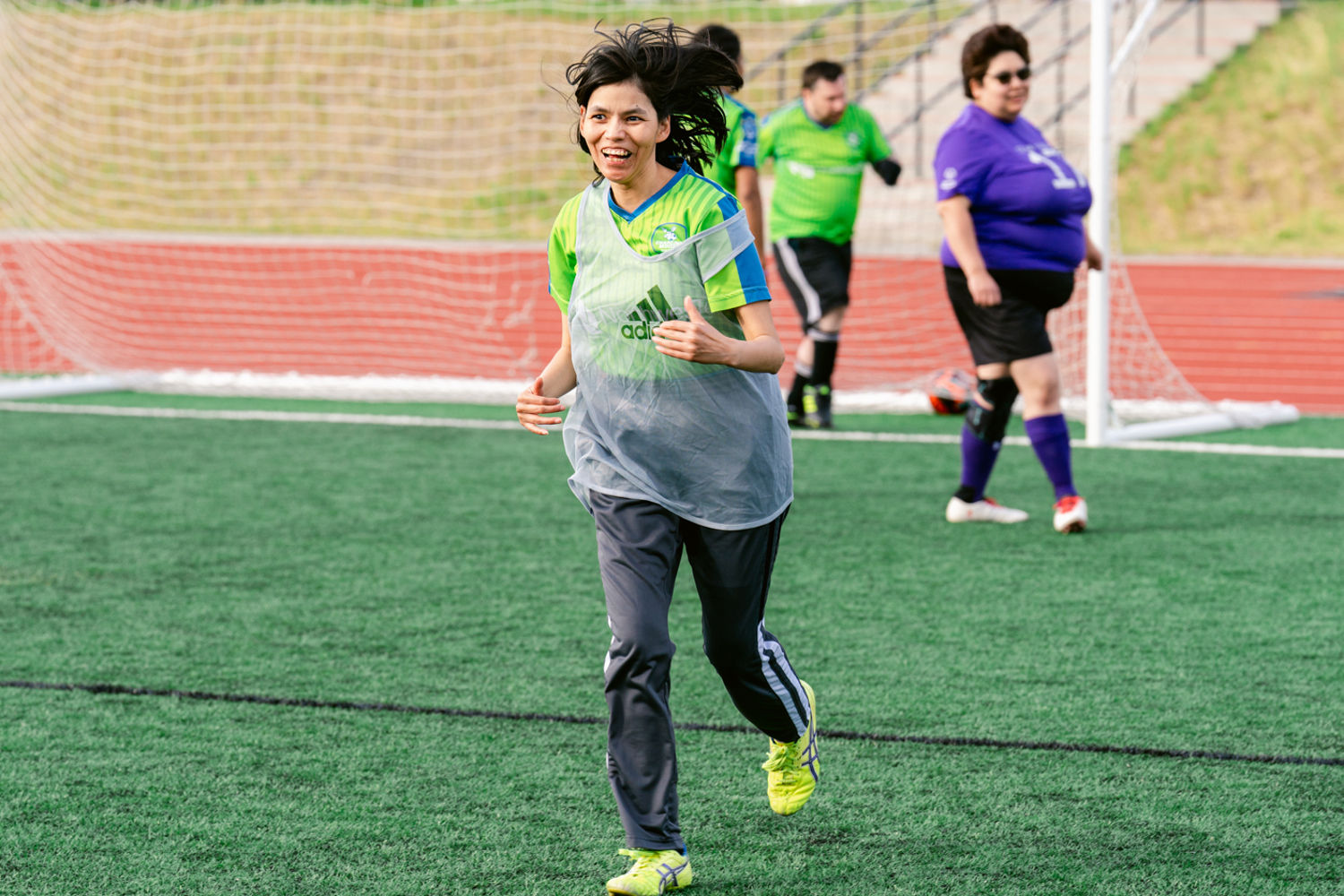 A female soccer player happily running on the soccer field. She is wearing a bright green jersey with a clear grey pinnie over top of it. She is also wearing bright yellow soccer cleats. Behind her is the soccer net and three other soccer players out of focus.