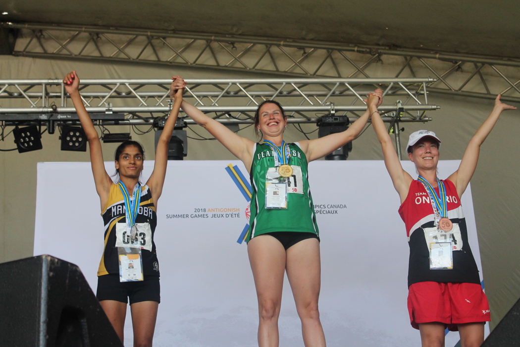 Three Special Olympics athletes standing on the podium. Saskatchewan's athlete in the middle has won gold. Manitoba's athlete on the left has won silver. Ontario's athlete on the right has won bronze. All three athletes are holding hands and holding them up in the winners position.