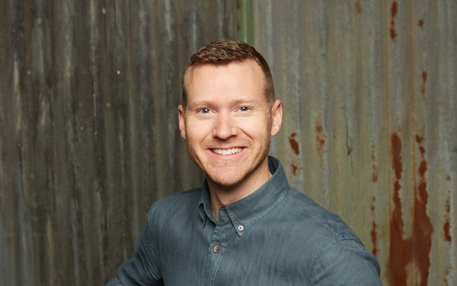 Headshot of a man wearing a grey button up shirt and beige pants.