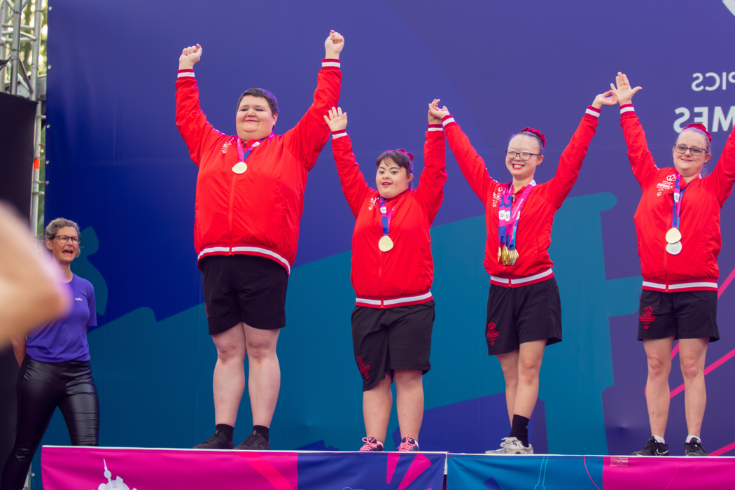 Diverse group of people in red outfits stand on a podium. All have medals on neck and arms in the air.  
