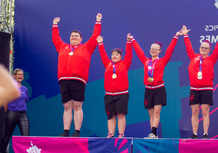 Diverse group of people in red outfits stand on a podium. All have medals on neck and arms in the air.