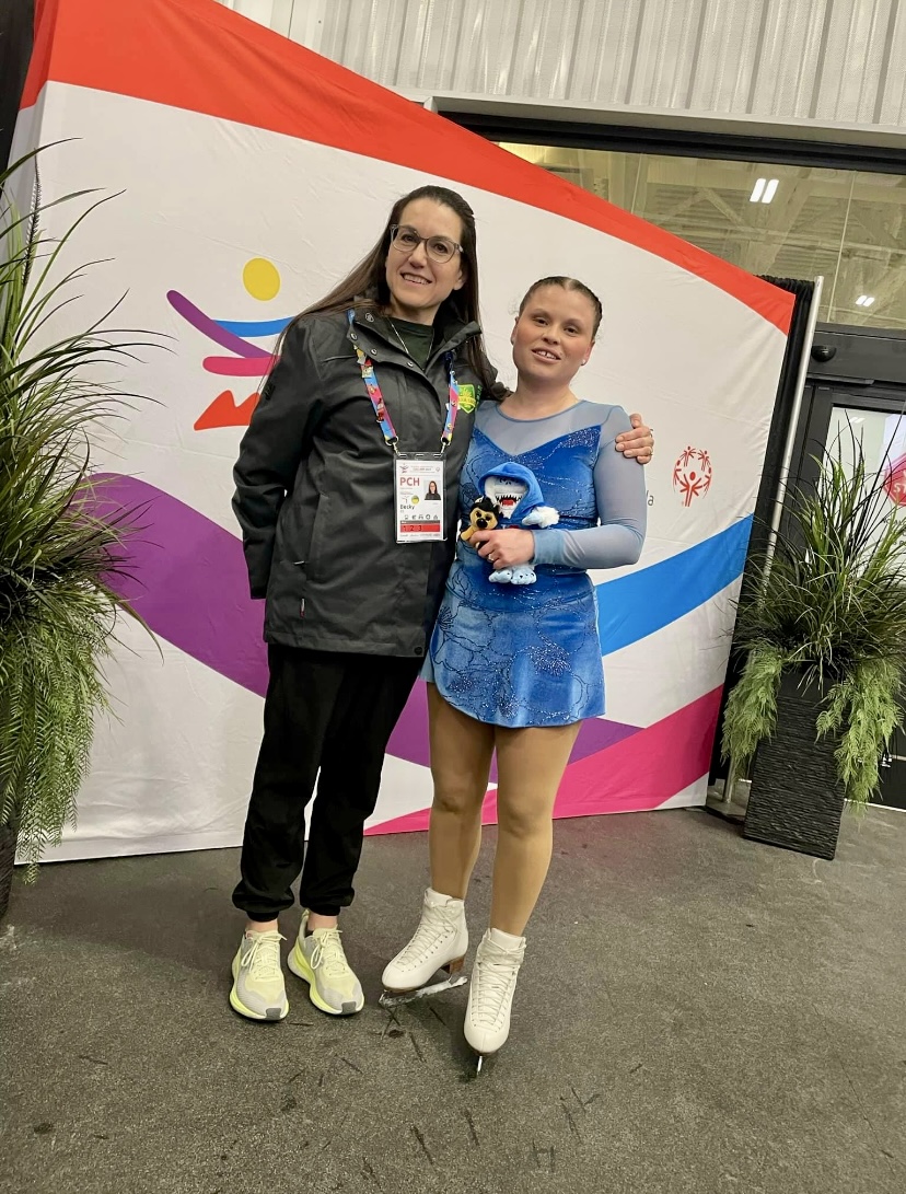 A young figure skating athlete is posing with her coach. They are both smiling for the camera and standing in front of a backdrop. The figure skater is still in her skates and blue figure skating outfit. The coach is wearing black pants and a black jacket with a colourful lanyard hanging around her neck. A young figure skating athlete is posing with her coach. They are both smiling for the camera and standing in front of a backdrop. The figure skater is still in her skates and blue figure skating outfit. The coach is wearing black pants and a black jacket with a colourful lanyard hanging around her neck.