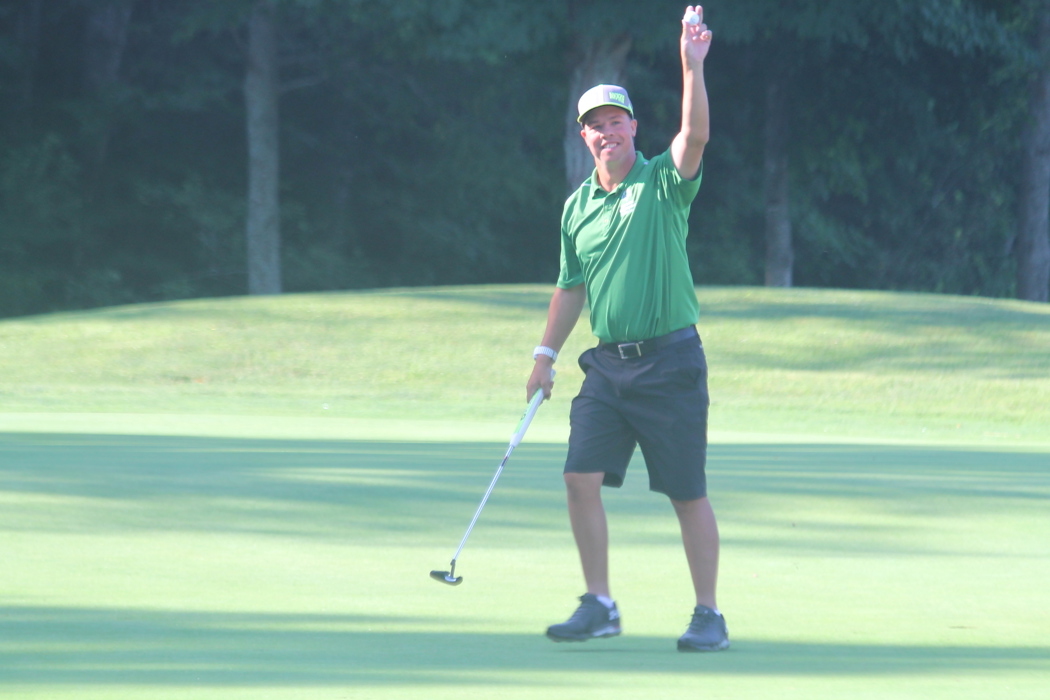 A golf athlete is standing on the golf course holding up a golf ball in one hand. In the other hand, he is holding his golf club. He is smiling for the camera and wearing a green golf shirt, grey shorts, and a grey baseball cap.