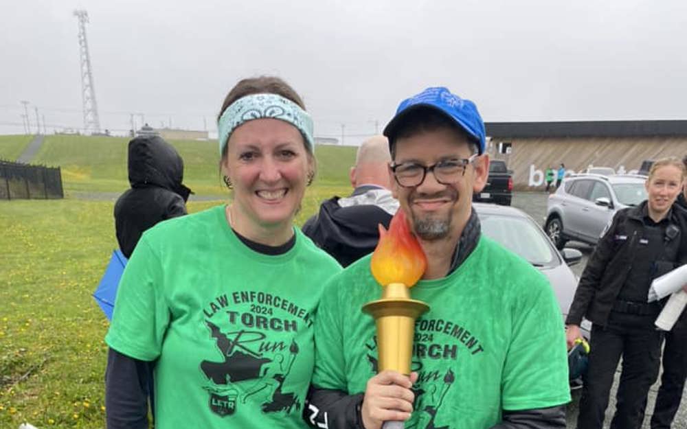 Two people standing beside each other, both in green law enforcement torch run shirts. One is holding the torch. 