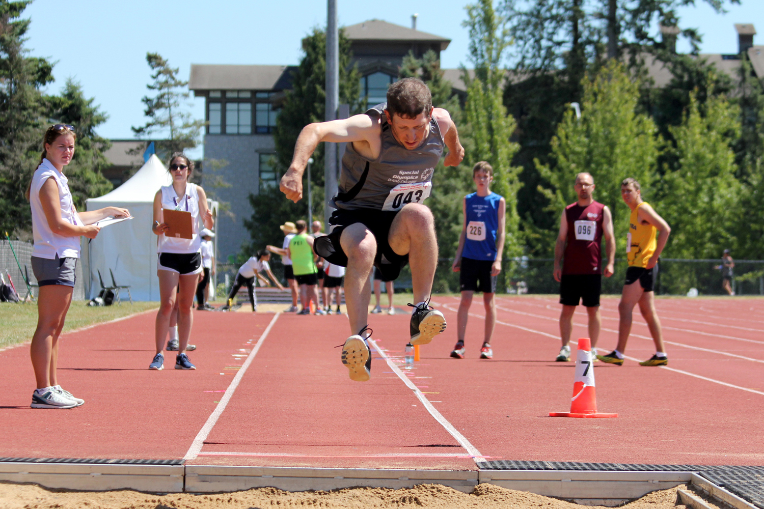 Special Olympics BC athlete competing in long jump is shown mid-jump before landing in the sand.