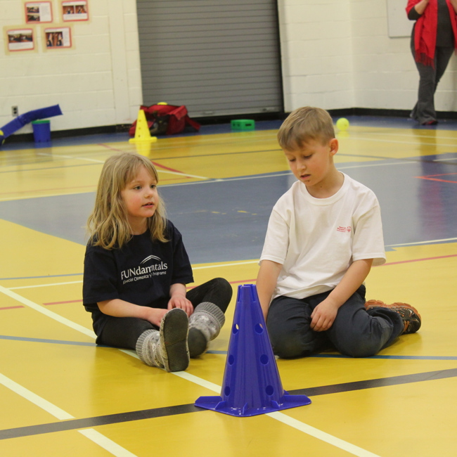 Two small children are sitting on a gym floor in front of a blue pylon. The girl on the left is wearing a black FUNdamentals t-shirt and the boy on the right is wearing a white Special Olympics Saskatchewan t-shirt.