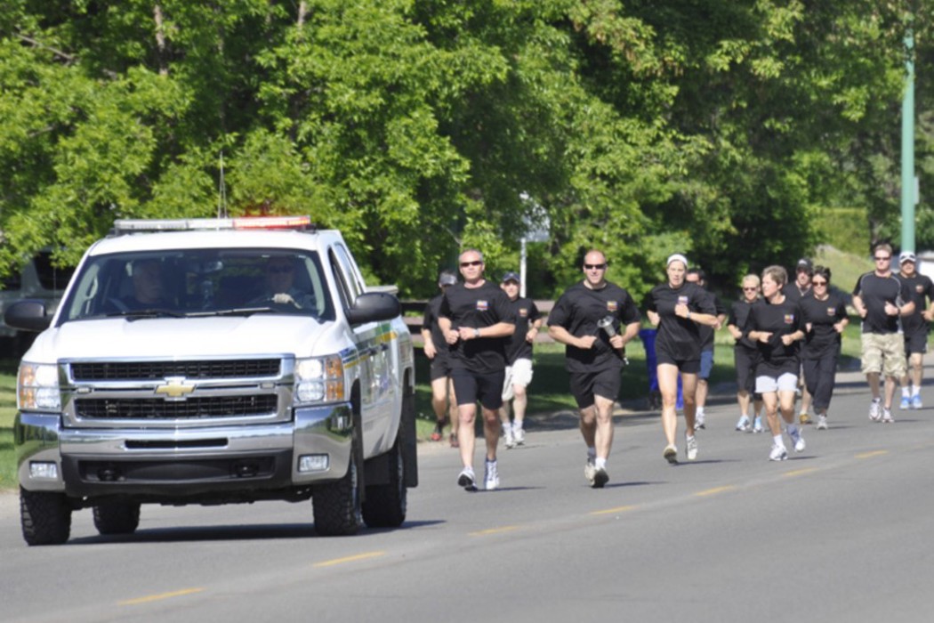 A group of Law Enforcement Torch runners can be seen running in the distance. They are all wearing black t-shirts and some are wearing sunglasses. They are running next to a white police truck with it's lights on.