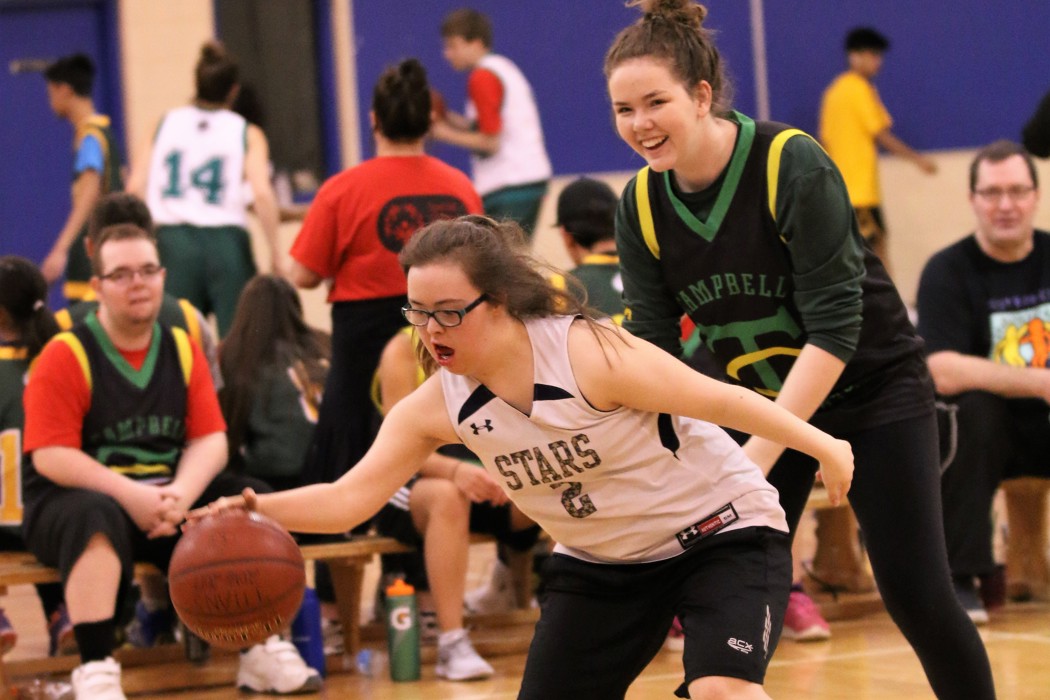 Action shot photo of two athletes playing basketball. One athlete in a white jersey has possession of the basketball. The other athlete is standing behind her and wearing a black jersey. Behind the play, you can see other athletes sitting on the bench.