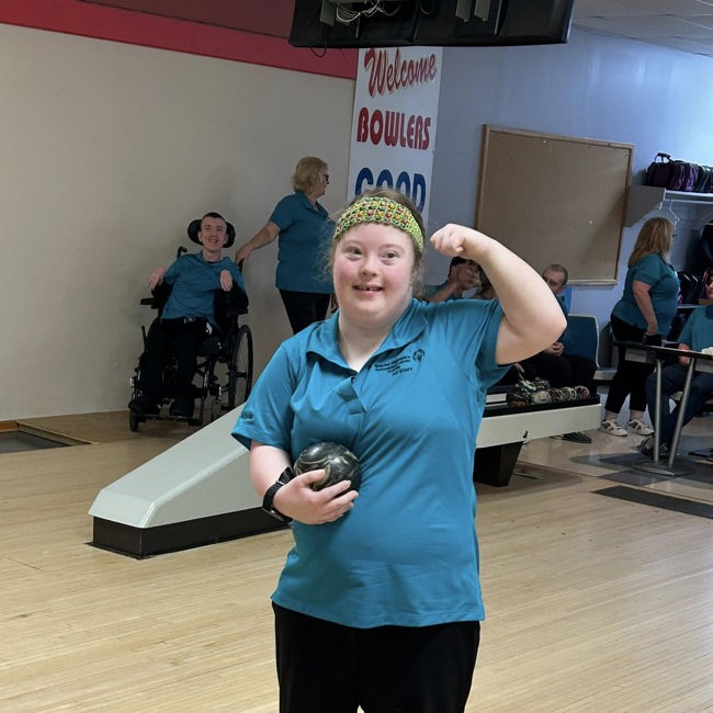An athlete is standing at the start of the bowling aisle. She is holding a bowling ball in one arm while the other arm is raised to show off her muscles. She is wearing a blue bowling t-shirt and smiling for the camera.