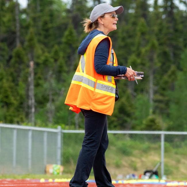 A coach, on the sideline, watching the match with her clipboard ready to take notes on what she is watching. She is wearing a high visibility vest, a baseball cap, and sunglasses.