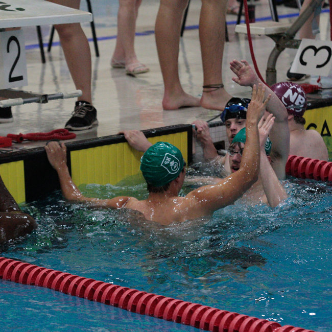 Six swimmers are holding on to the side of the pool, in the water, and giving each other high-fives. The two athletes in the front of the image have their backs turned towards the camera while you can just see the faces of the two athletes they are high-fiving.