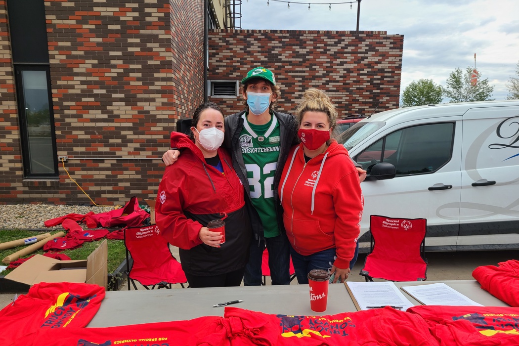 Three fundraisers preparing for an event. One individual in the middle is wearing a green Saskatchewan jersey, while the other two individuals beside him are wearing a red jackets/sweater. They are standing behind a table that has many red t-shirts on it. The three fundraisers are wearing masks.