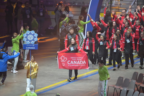 Group of athletes walking into opening ceremony. Front athlete is holding a Canadian banner with the words Special Olympics Team Canada. All athletes are dressed in red and black. 
