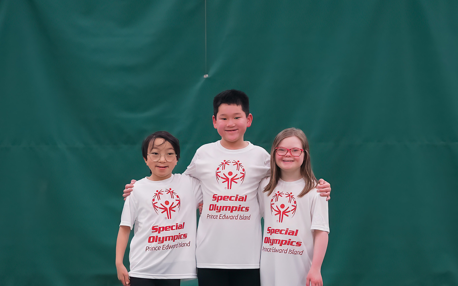 Three young kids stand in a line. All wearing Special Olympics white shirts and black pants. 