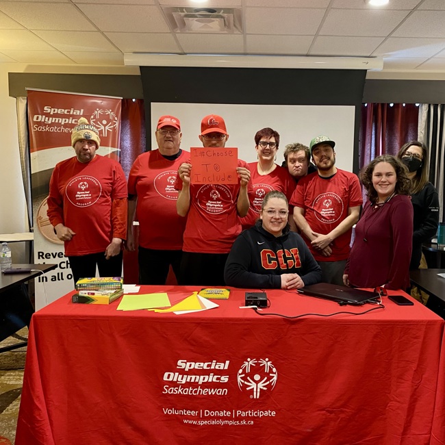 A group of athletes and volunteers are sitting and standing behind a table and smiling for the camera. Many people are wearing red Special Olympics Saskatchewan long sleeved shirts.