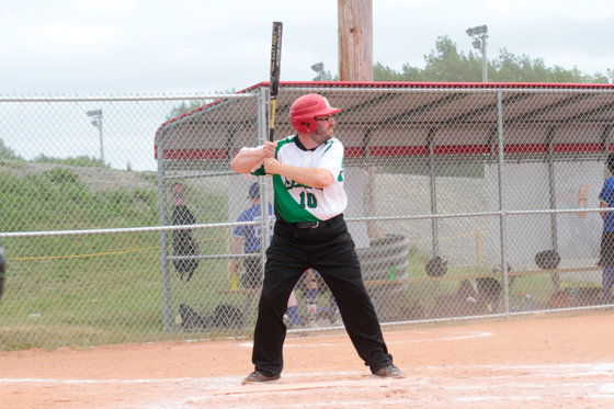 A baseball athlete at the base getting ready to hit the baseball. They are wearing a green Special Olympics Saskatchewan jersey with a red helmet. The baseball bat is swung behind their shoulder.