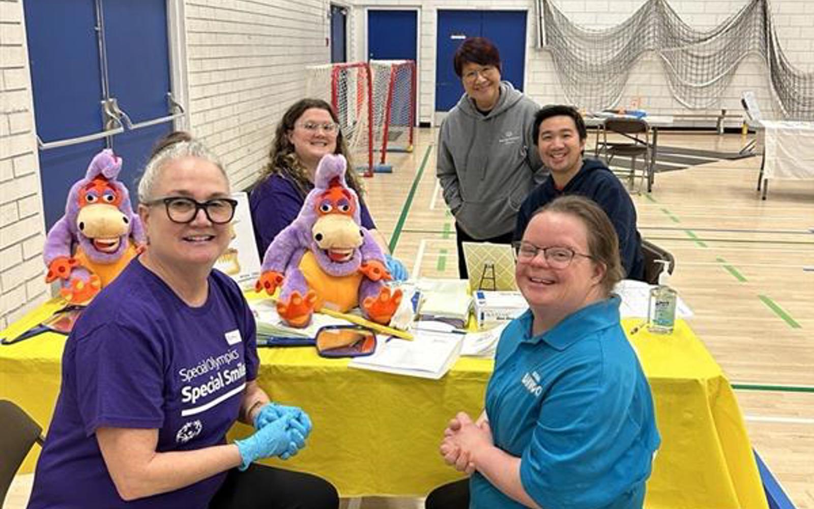 A group of athletes and volunteers sit around a table and smile at the camera.