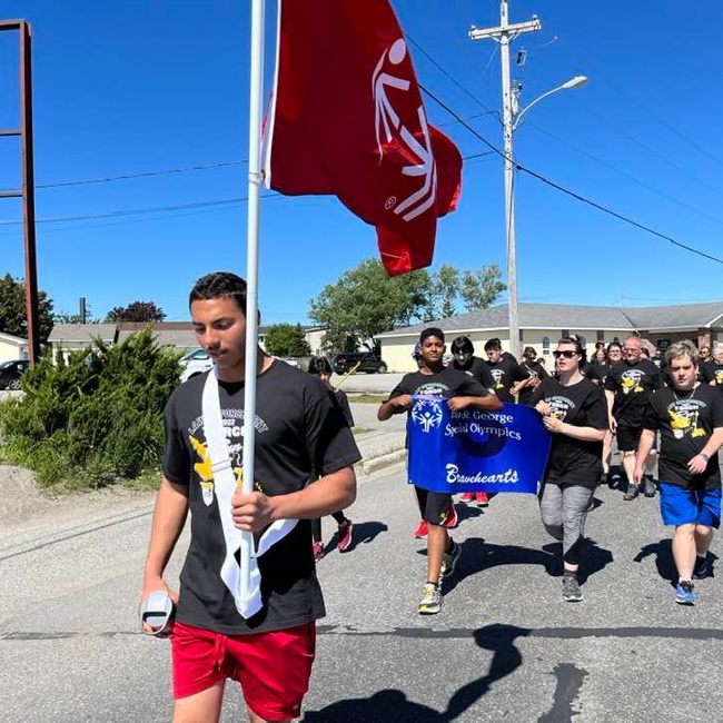 Law Enforcement Torch Runners are walking down the street in matching Law Enforcement Torch Run t-shirts. The leader of the group is holding up a large, red, Special Olympics flag. The people behind him are holding a "Bay St. George Special Olympics Bravehearts" banner.