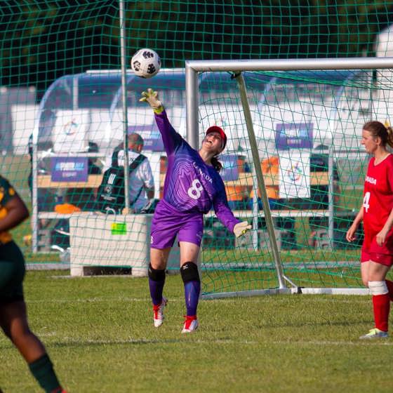 A soccer goalie standing in front of her soccer net, throwing the ball out onto the field. She is wearing a purple long sleeved jersey with Canada and the number 8 written on the front of it.