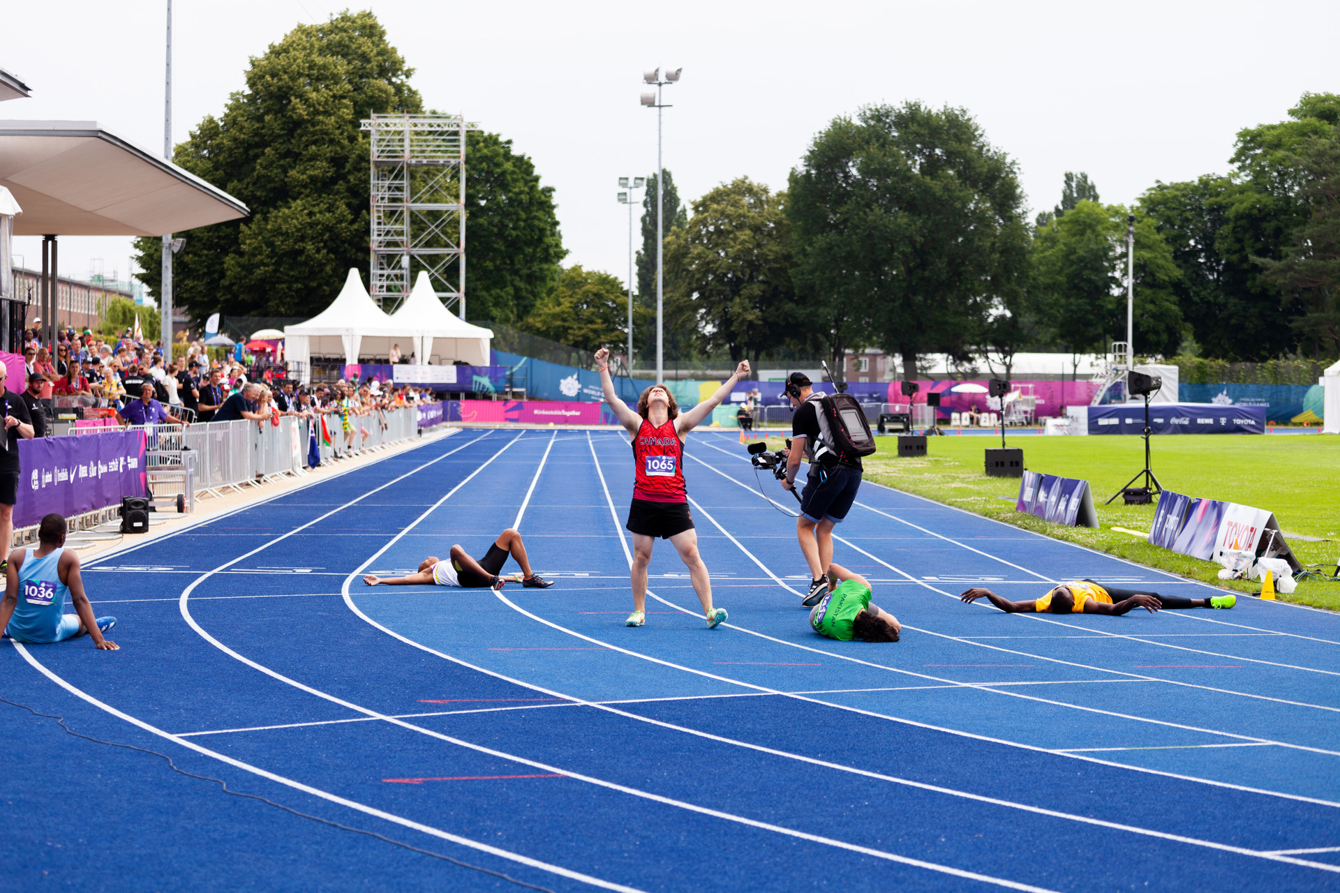 A triumphant Special Olympics BC athlete at the end of their race with their arms raised as fellow competitors lie exhausted on the track around him. 