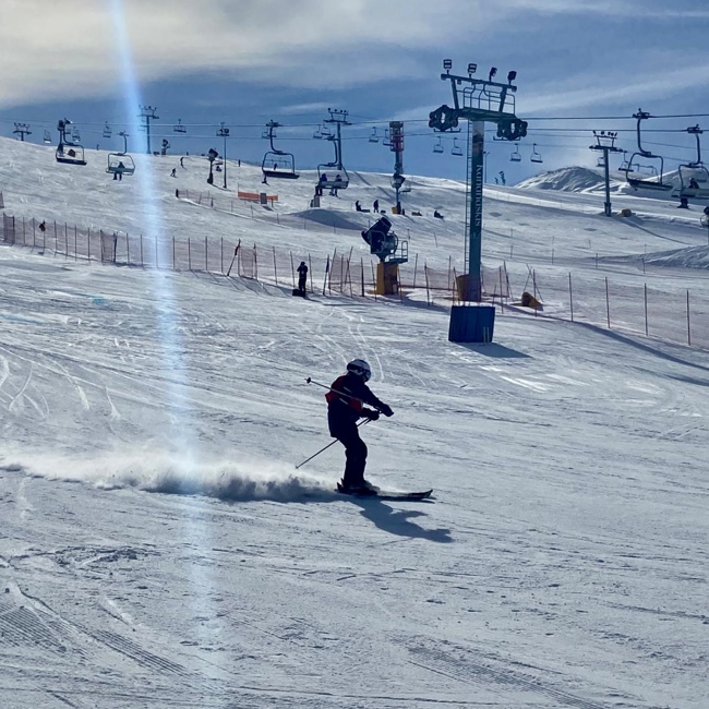 An action shot photo of an alpine skier going down the hill. The skier is a medium distance away from the camera and you can see the snow blowing behind their skis. In the distance behind the skier, you can see the ski lift and other skiers going down a different hill.