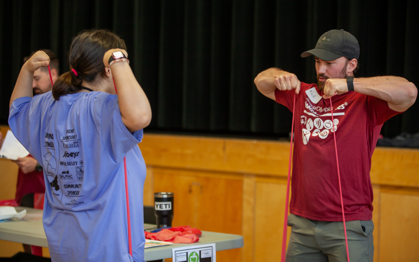 A volunteer instructor, is demonstrating and showing an athlete a stretching exercise using a red band. The athlete is following along with the instructor, with her back to the camera.