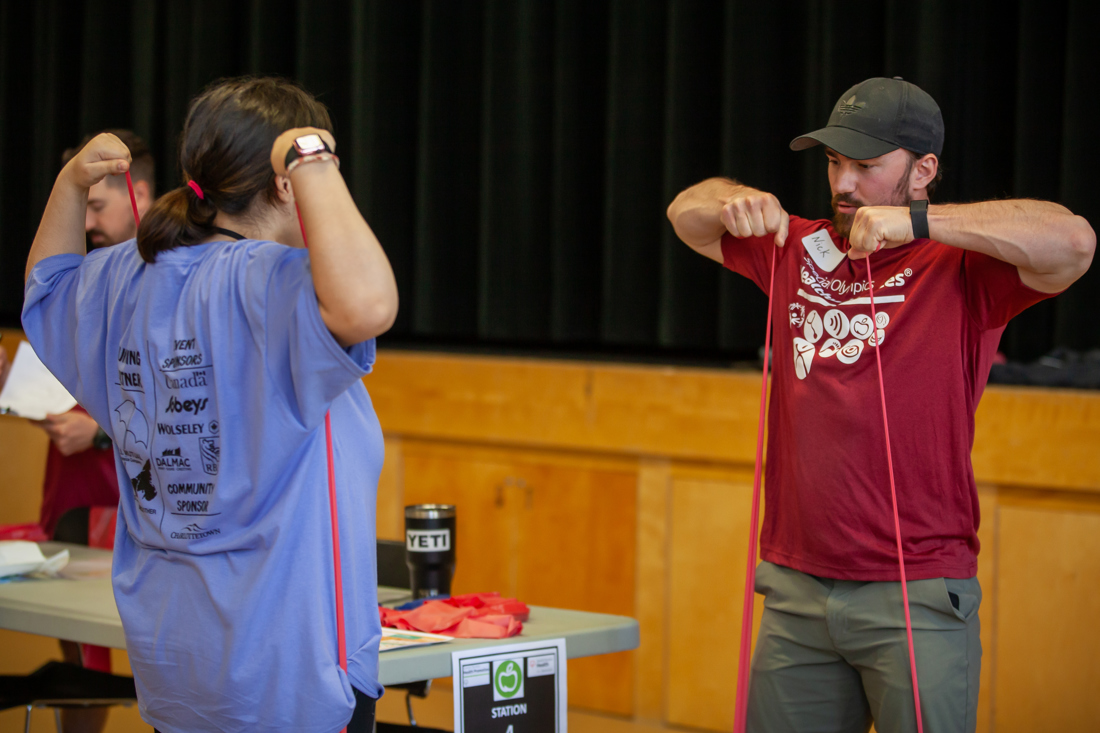 A volunteer instructor, is demonstrating and showing an athlete a stretching exercise using a red band. The athlete is following along with the instructor, with her back to the camera.
