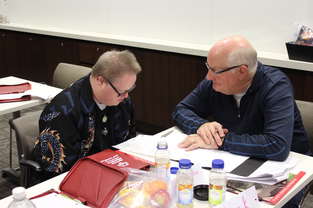 Two individuals are sitting at a table covered with papers and snacks. The individual on the right is reading something off the paper to the individual on the left.