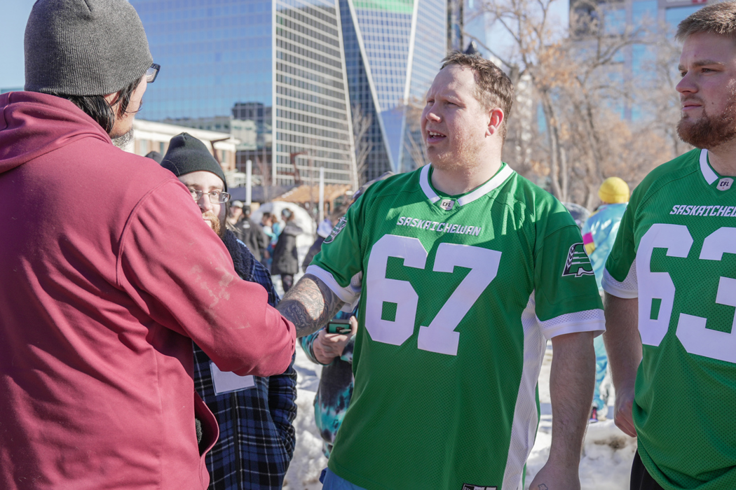 Two individuals are shaking each other's hand. One individual on the left has his back to the camera. The other individual is wearing a green "Saskatchewan" jersey with the number 67 on it and is looking at the individual he is shaking hands with..