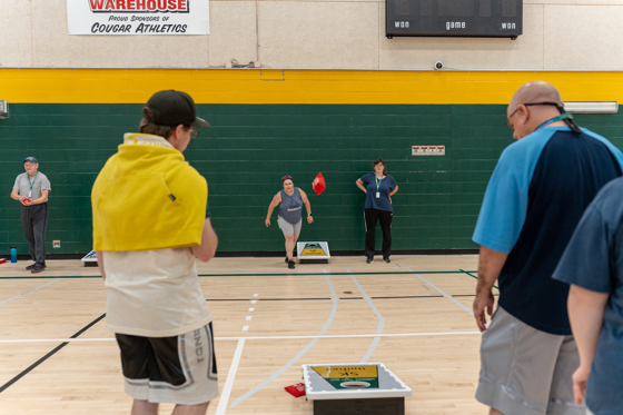 An action shot photo of people playing bean bag toss. In the distance you can see someone throwing a bean bag. The bean bag can be seen in midair coming closer to the camera. On the floor are the bean bag boards where the athletes are trying to toss the bean bags into. The game is being played inside a gymnasium.