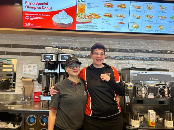 A female Special Olympics Saskatchewan athlete poses for the camera with a Tim Hortons employee. They are behind the Tim Hortons counter with their arms wrapped behind each other and they are smiling. Behind them are the display screens as well as coffee makers and equipment.