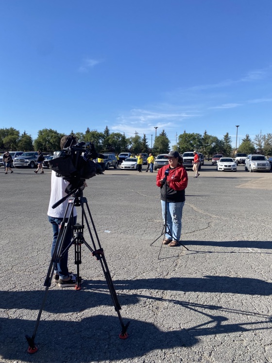 A medium shot photo of a camera on a tripod. There is an individual behind the camera, but the camera is obstructing him from view. There is also an individual in front of the camera. You can see their full body and they are wearing a red jacket with jeans.