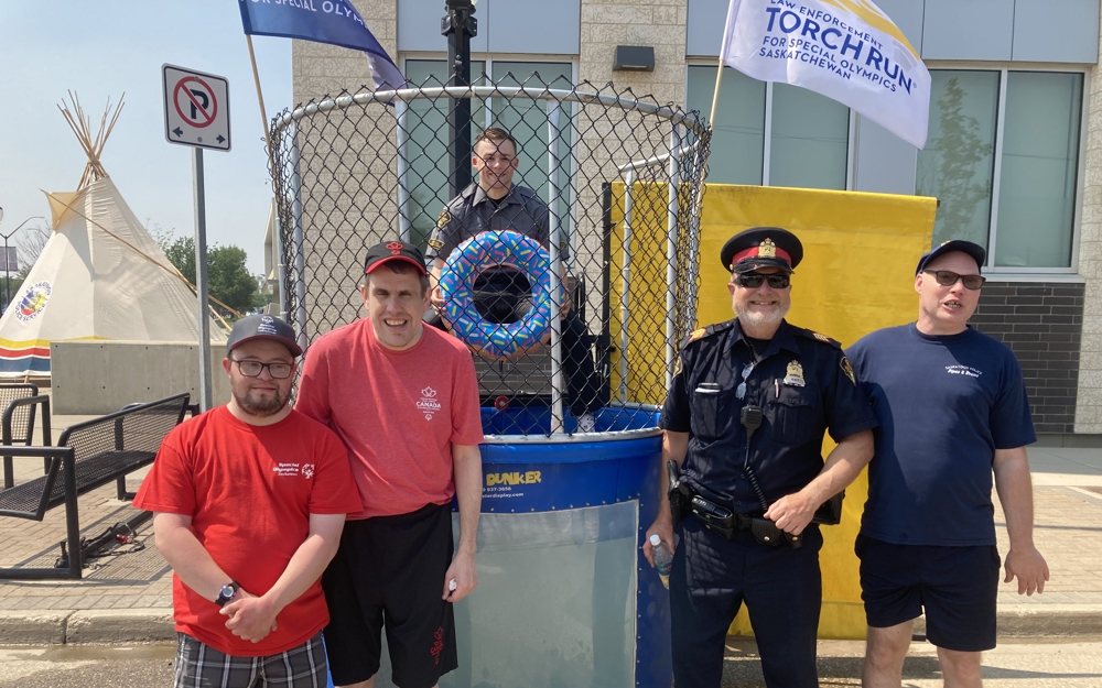 Three Special Olympics athletes are smiling and posing for the camera alongside two police officers. They are standing in front of a dunk tank and one of the police officers is inside the dunk tank.
