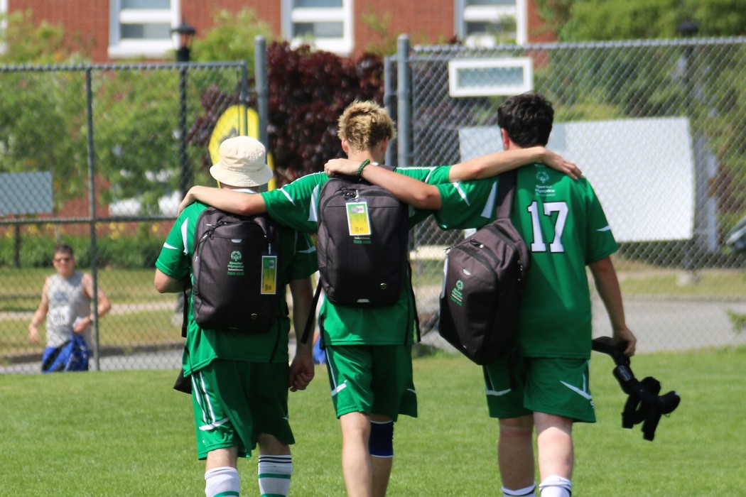 Three Special Olympics Saskatchewan soccer players, are walking off the field with their arms on each others shoulders. They are walking away from the camera so we can only see their backs. They are wearing their all green soccer uniform and have black backpacks on.