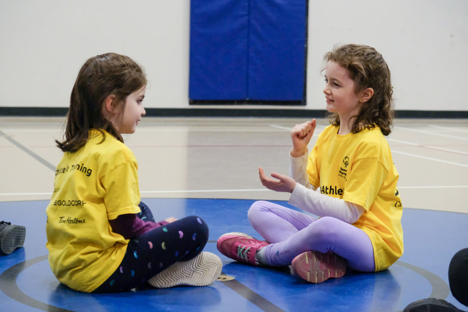 Two young girls with intellectual disabilities sit together wearing matching yellow shirts and playing a game with their hands.