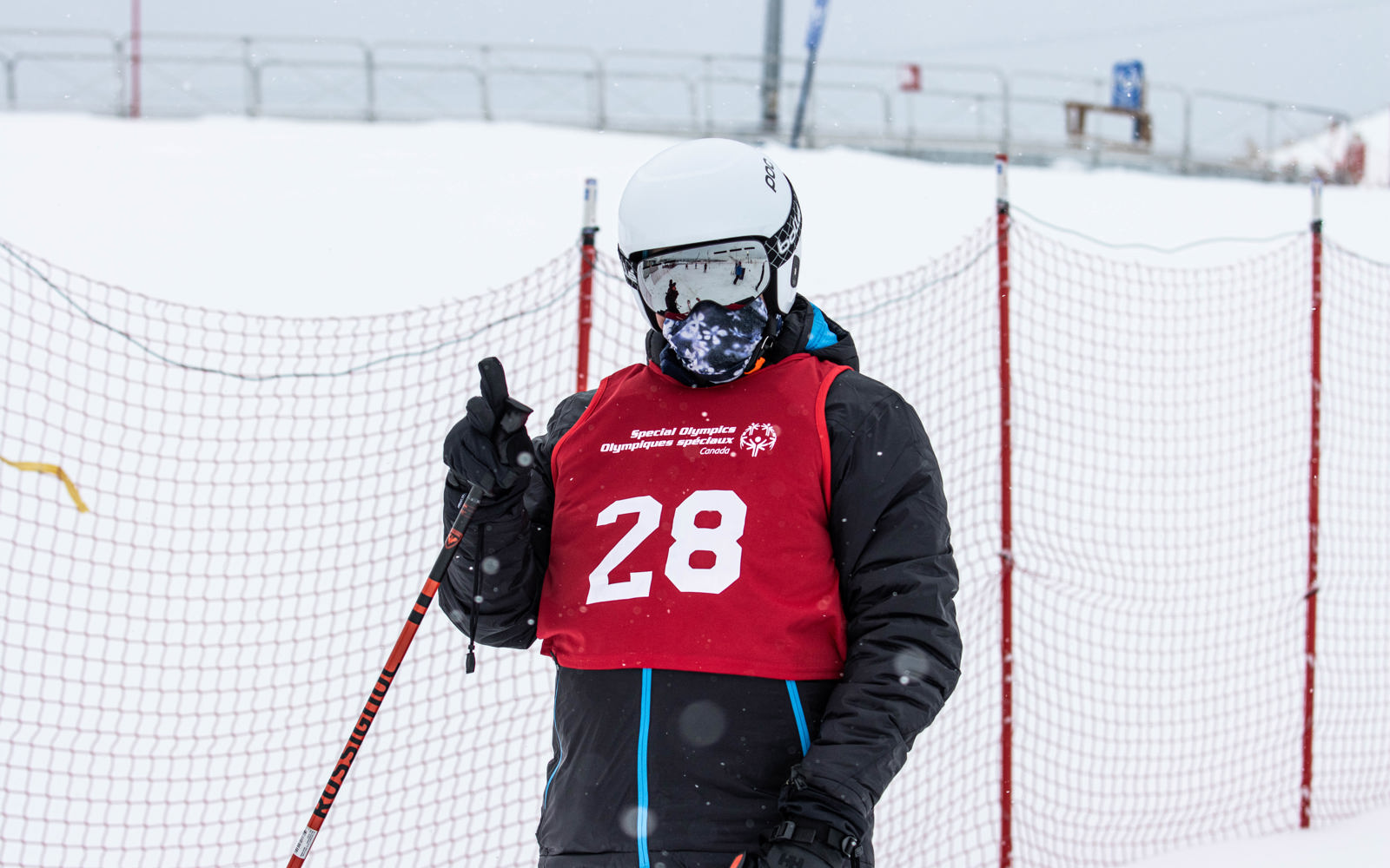 Special Olympics BC alpine skiing athlete wearing a helmet, goggles, and red racing bib gives the camera a thumbs up.