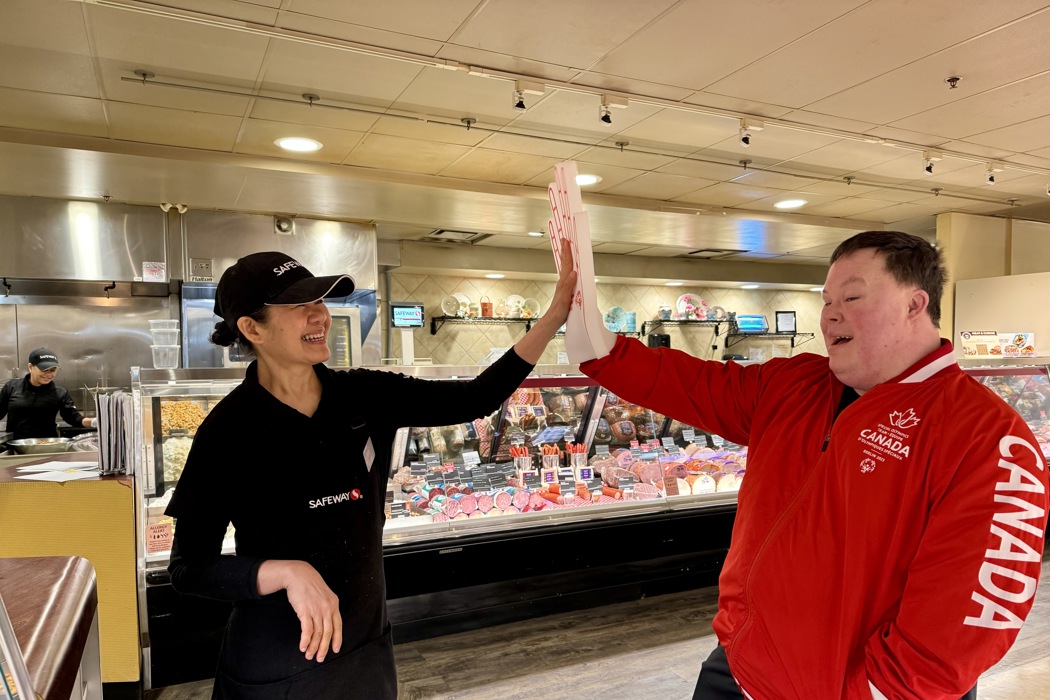 A Special Olympics Saskatchewan athlete is high-fiving a Safeway employee. They are both smiling and happy. The Safeway employee is wearing her black uniform while the athlete is wearing a red Canada sweater and a white foam hand.