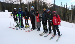 A group photo of the skiers at training camp. They are at the top of the hill and have their ski gear on. Everyone is looking towards the camera and smiling. You can see a large mountain in the background.