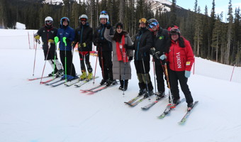 A group photo of the skiers at training camp. They are at the top of the hill and have their ski gear on. Everyone is looking towards the camera and smiling. You can see a large mountain in the background.