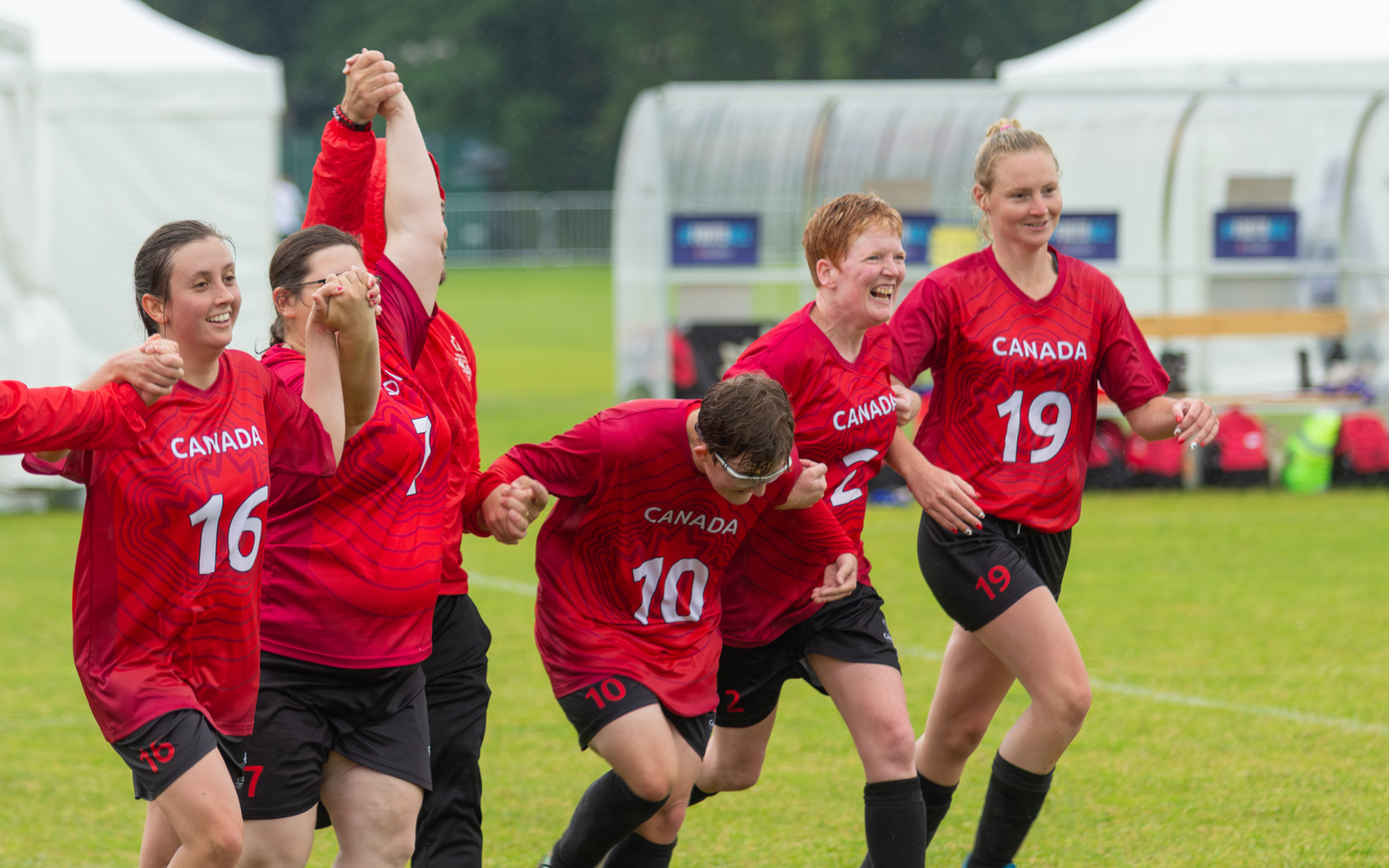 6 female athletes dressed in red Canada soccer jerseys and black shorts holding hands, smiling and coming toward the camera. All look like they just participated in a soccer match.