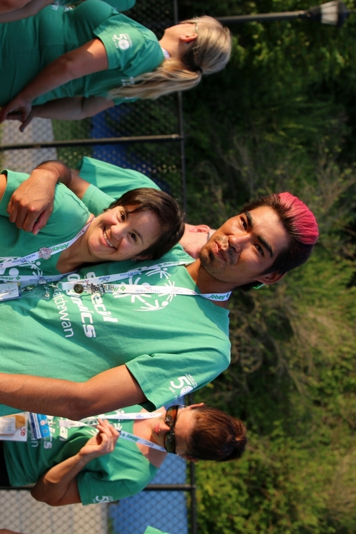 Two Special Olympics Saskatchewan athletes are wearing matching green "Special Olympics Saskatchewan" t-shirts and smiling for the camera. They have their arms wrapped behind each other and they are standing on a track.
