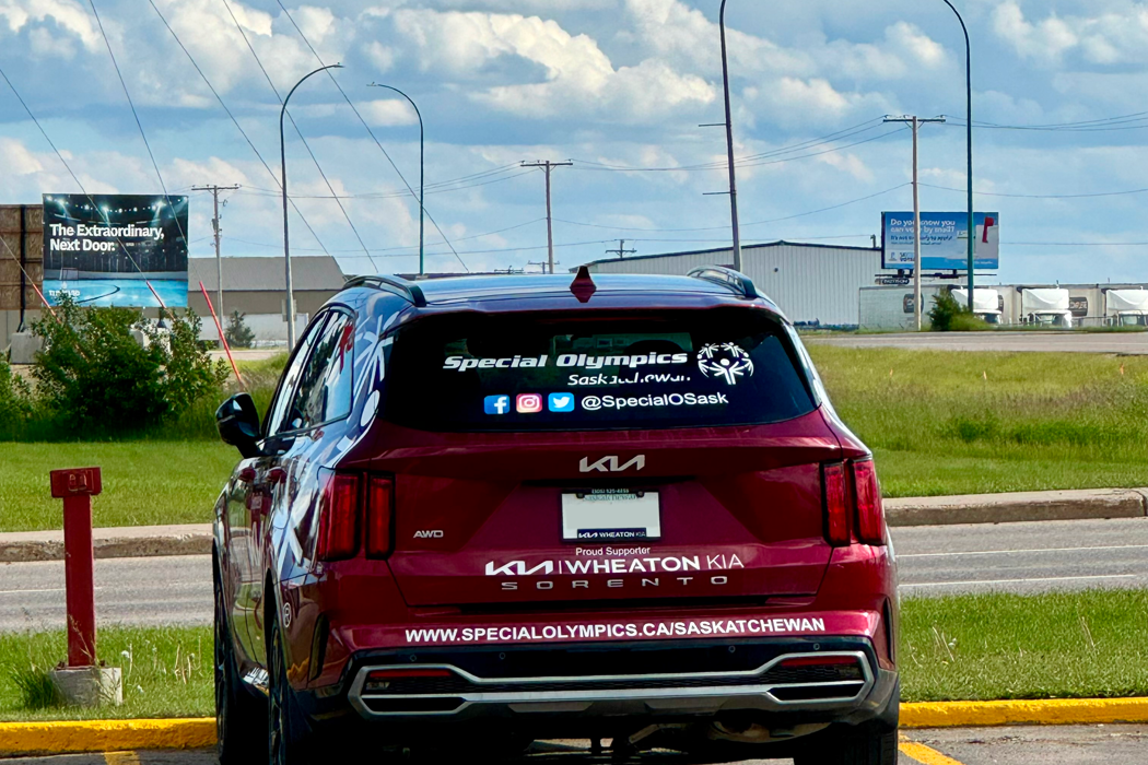 A red Kia car is parked in a parking spot. The car has the Special Olympics Saskatchewan logo on the window along with the social media icons and social media handle in white letters. Also in white letters and written along the bottom of the car is Special Olympics Saskatchewan's website.