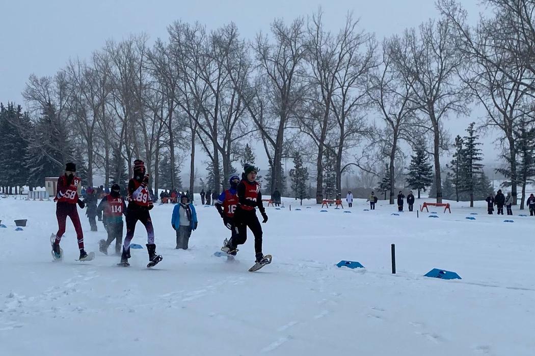 An action shot image of snowshoers. There are five athletes running across the snow a medium distance away from the camera. In the distance behind them, you can see trees and viewers watching the sport.