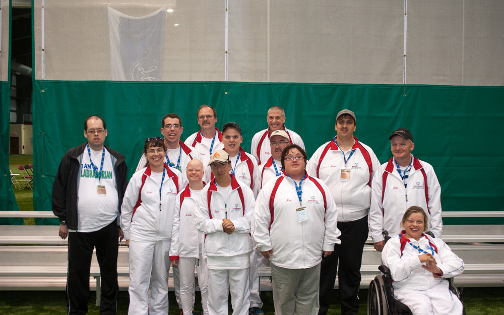 A team photo. Everyone is wearing white tracksuits and blue lanyards around their necks. They are standing in an indoor field.