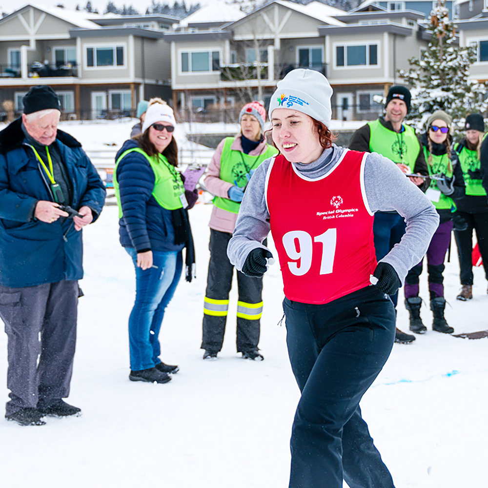 An athlete with intellectual disabilities wearing a red race bib participates in a snowshoeing race while volunteers and coaches watch.