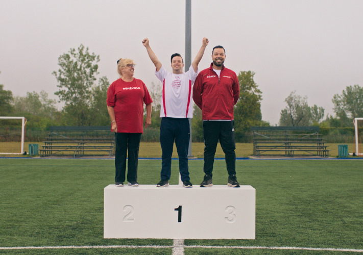 Three people stand on a podium in a soccer field. Woman on the left is on top of placement 2 and is wearing a red shirt. Man in middle is on top of placement 1 and has his arms in the air. Man of right is on top of placement 3 and is wearing a red sweater.