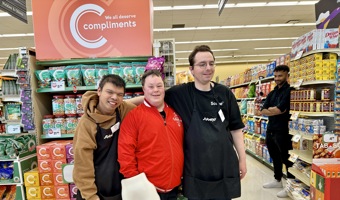 A Special Olympics athlete poses and smiles for the camera with two Sobeys workers. They are standing in a Sobeys grocery store, in front of one of the aisles. The Sobeys employees are wearing their black "Sobeys" aprons while the athlete is wearing a red sweater and a white foam hand.